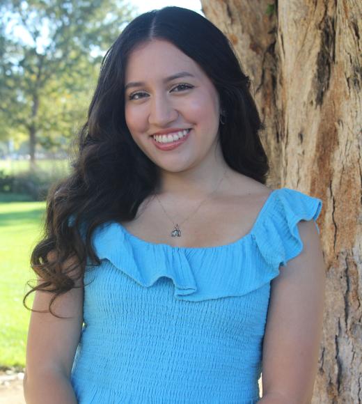 Tamara F standing is standing in front of a tree with a greenary background. 