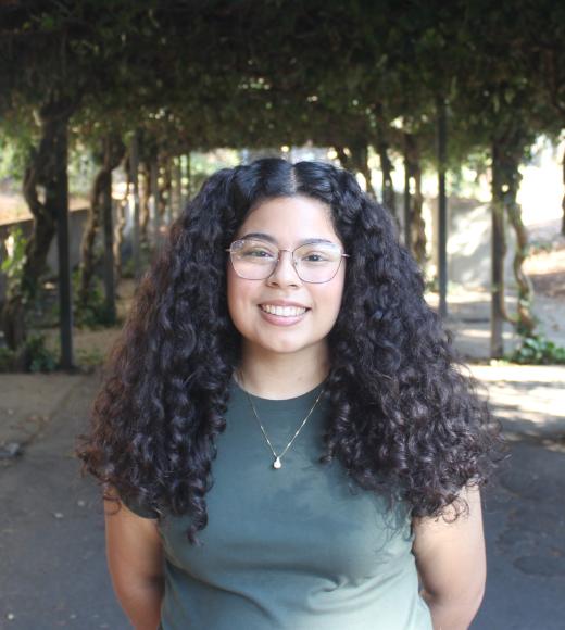  Izabel smiling warmly in front of greenery.