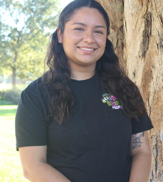 Xochitl is standing in front of a tree trunk, with greenary background.