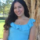 Tamara F standing is standing in front of a tree with a greenary background. 