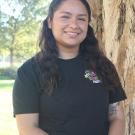 Xochitl is standing in front of a tree trunk, with greenary background. 