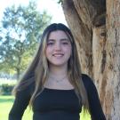 Lucia is standing in front of a tree trunk with greenary background,